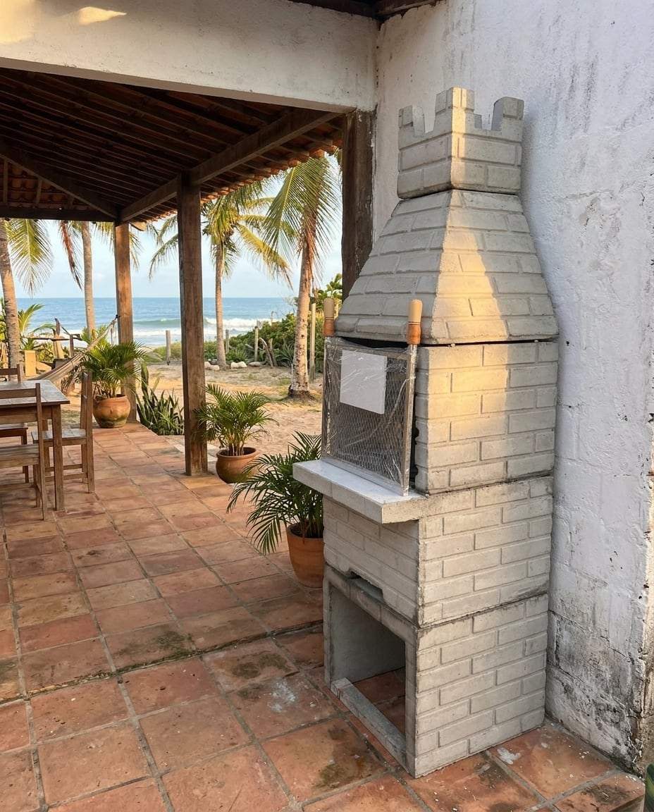 Simulação de ambiente: churrasqueira pré-moldada em cimento natural na varanda de uma casa de praia, com coqueiros e o mar ao fundo